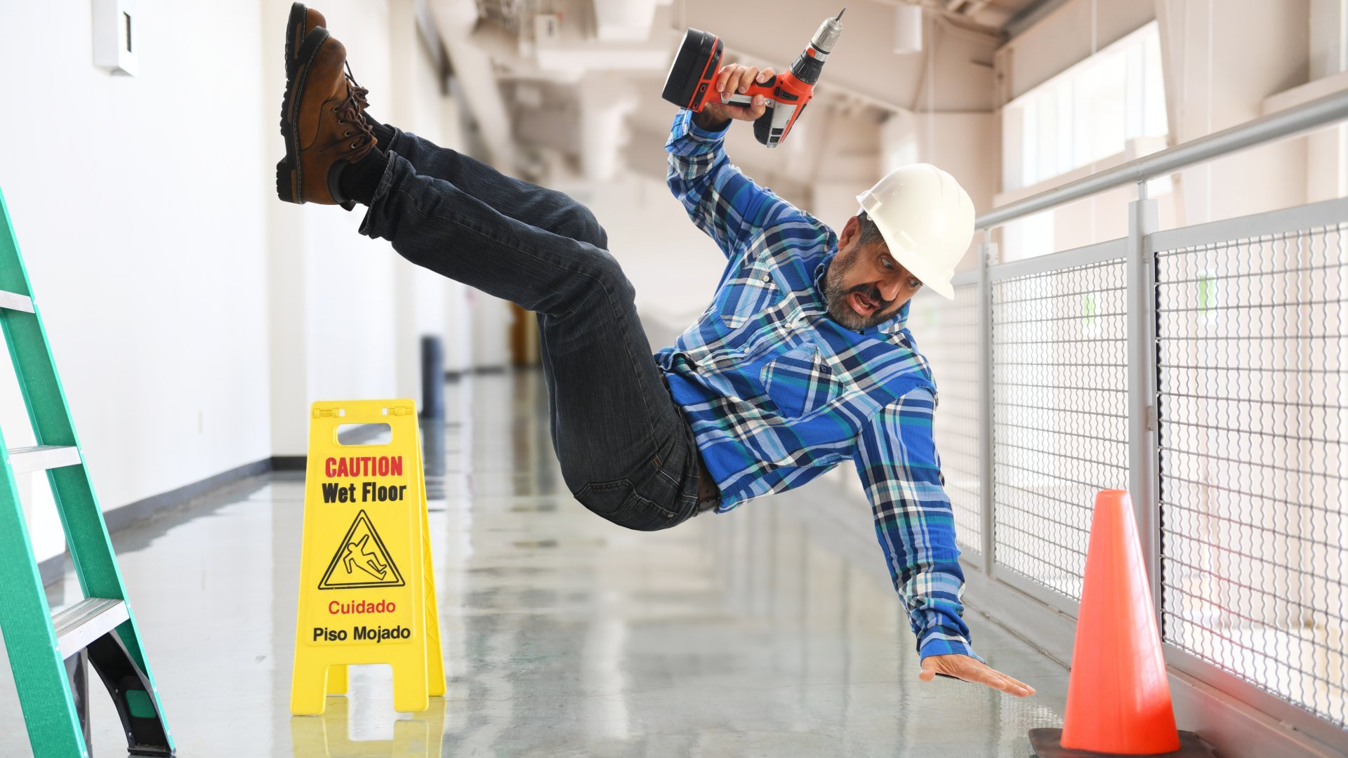 Construction worker falling, holding drill, near wet floor sign and safety cone.
