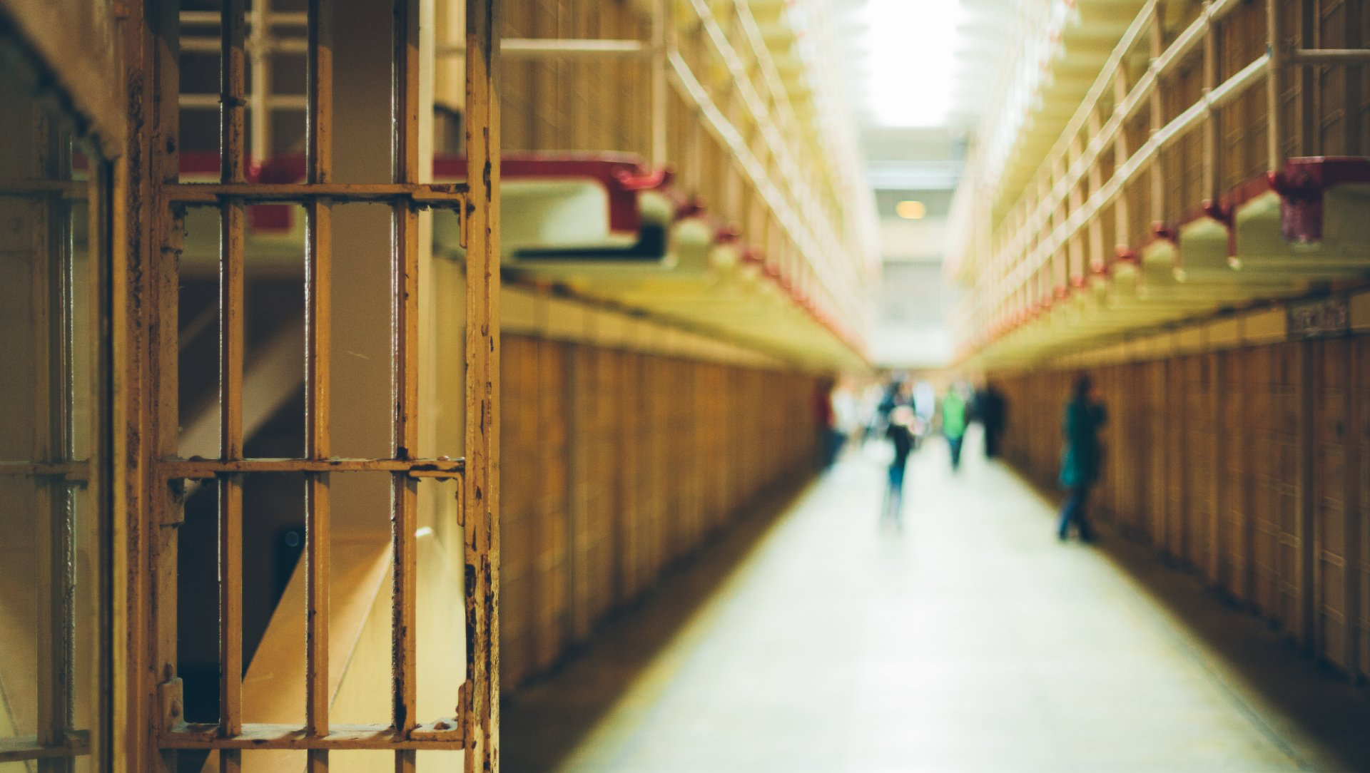 Prison cell bars in foreground, long hallway with people blurred in background.