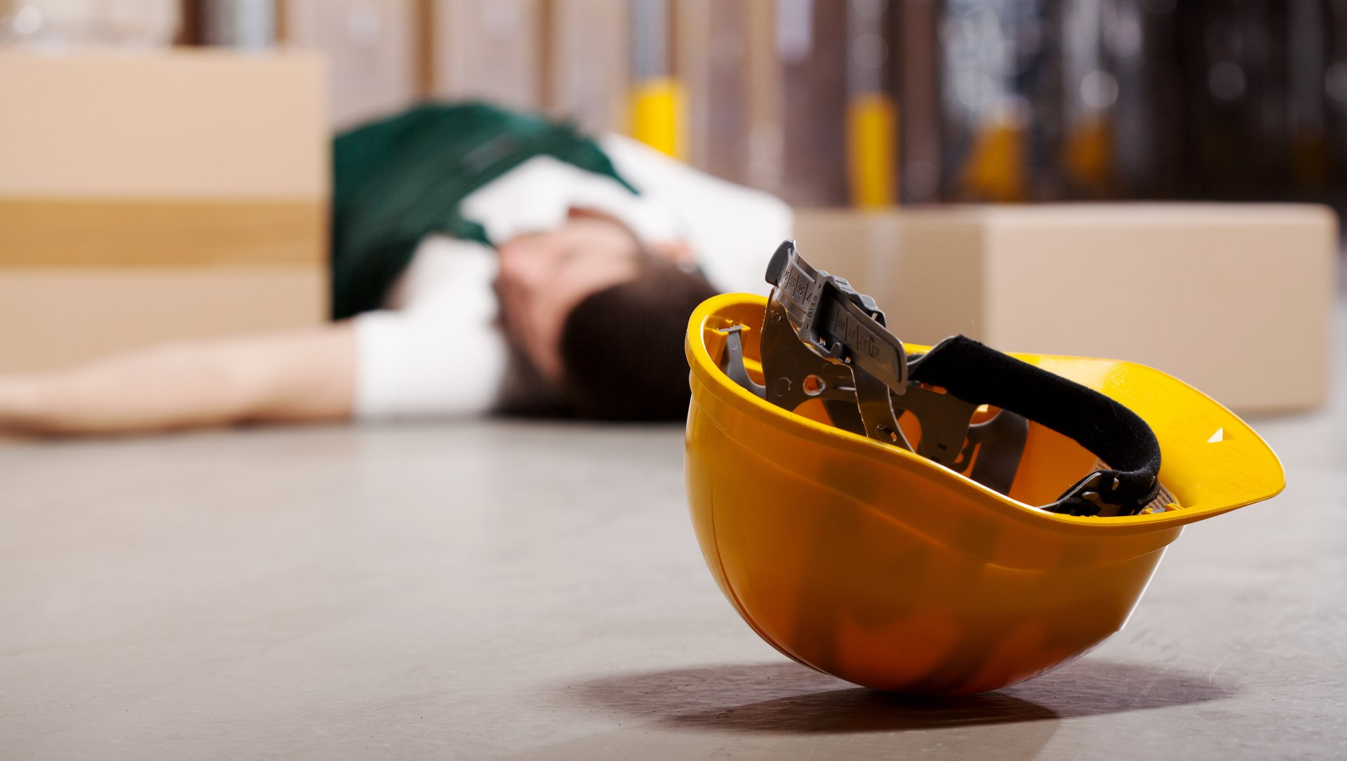 Yellow hard hat with glasses on the floor near a person lying down in a warehouse.
