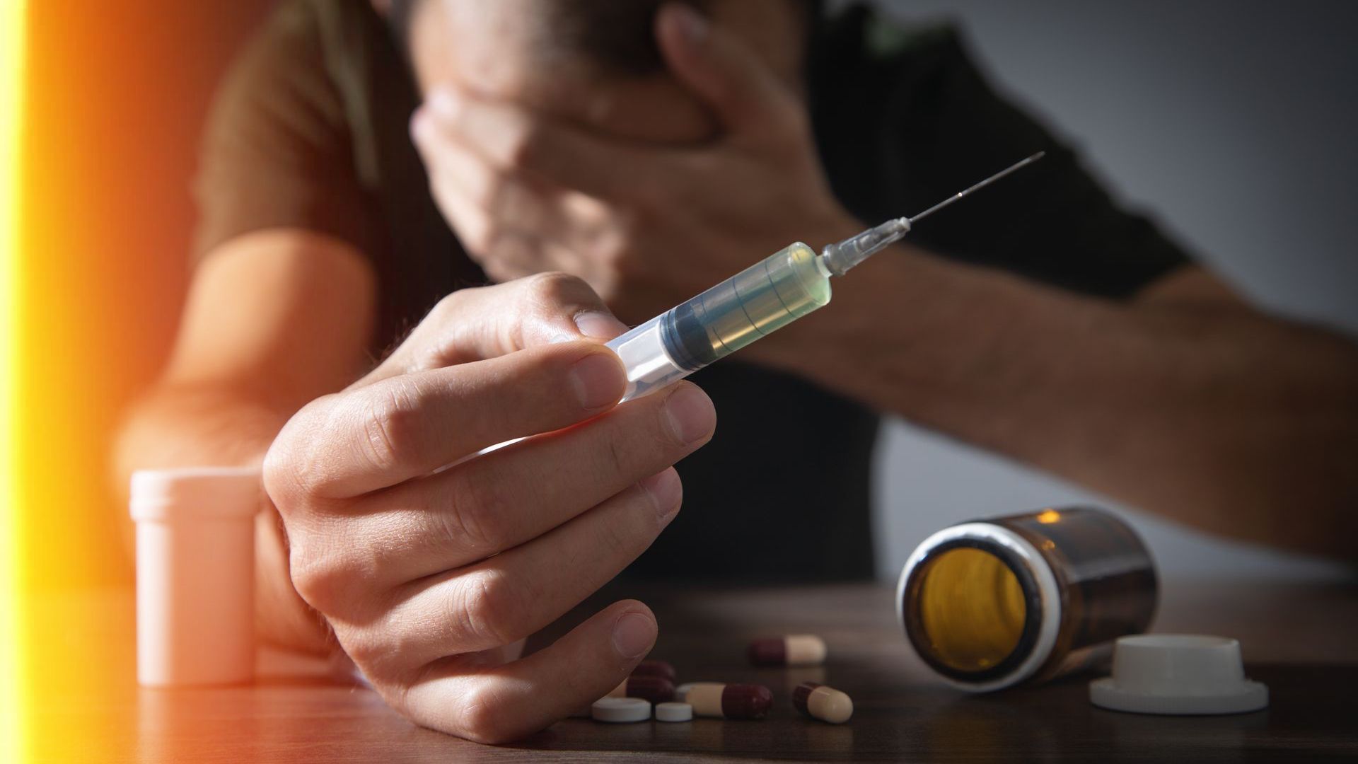 Person holding a syringe with liquid, pills, and bottle on a table. Person looks distressed.