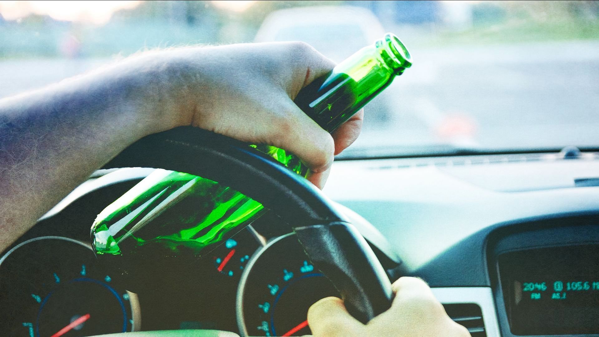Hand holding beer bottle on car steering wheel; dashboard visible.