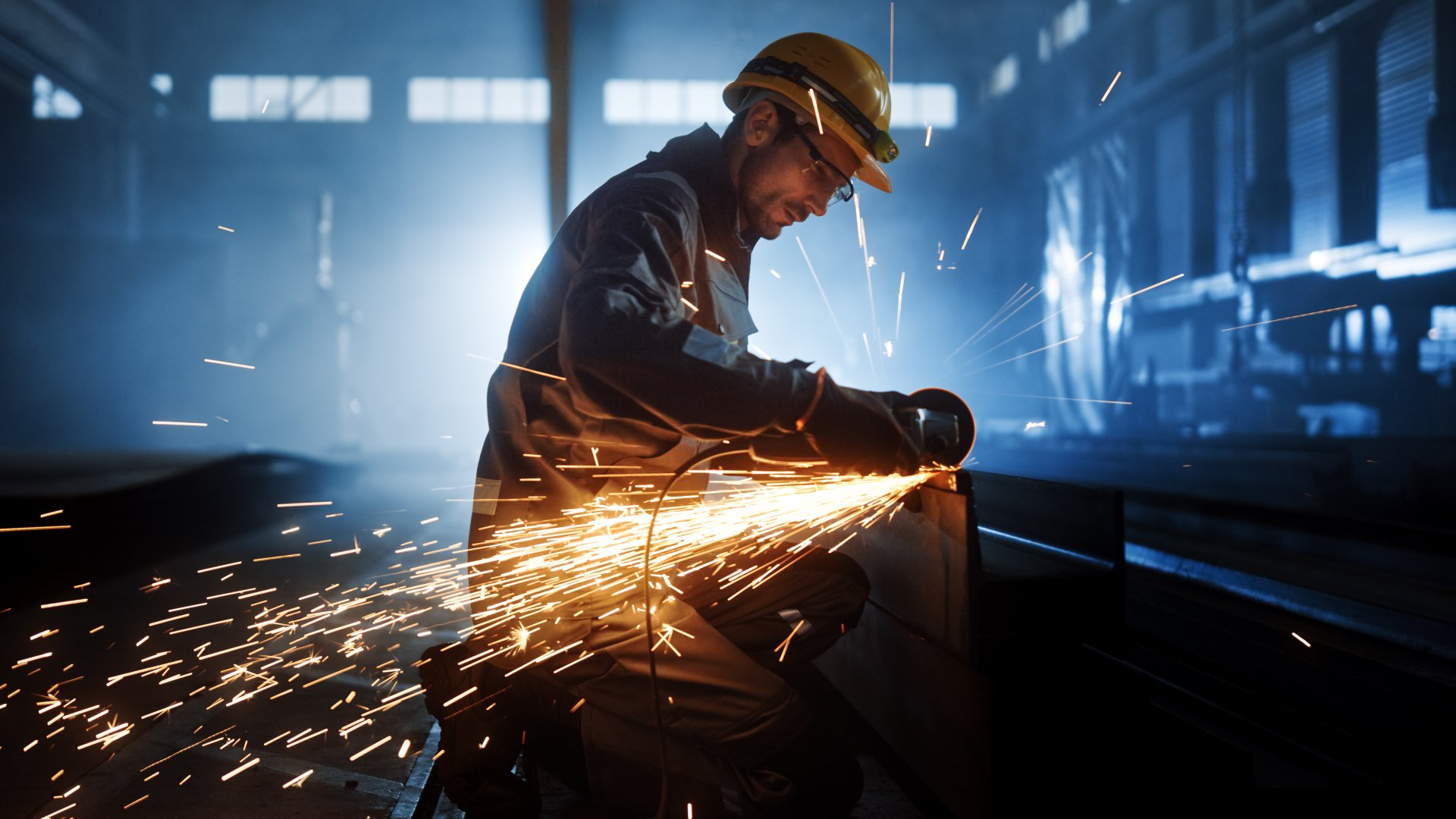 Worker wearing protective gear grinds metal, creating sparks in an industrial setting.