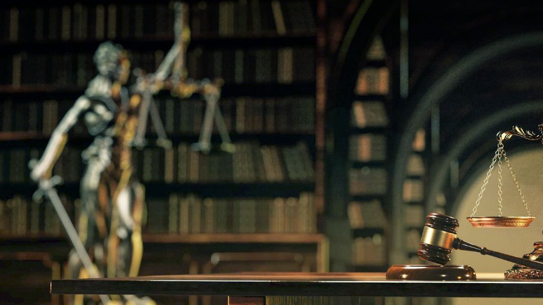 Blurred justice scale and gavel on desk with Lady Justice statue and bookshelf background.