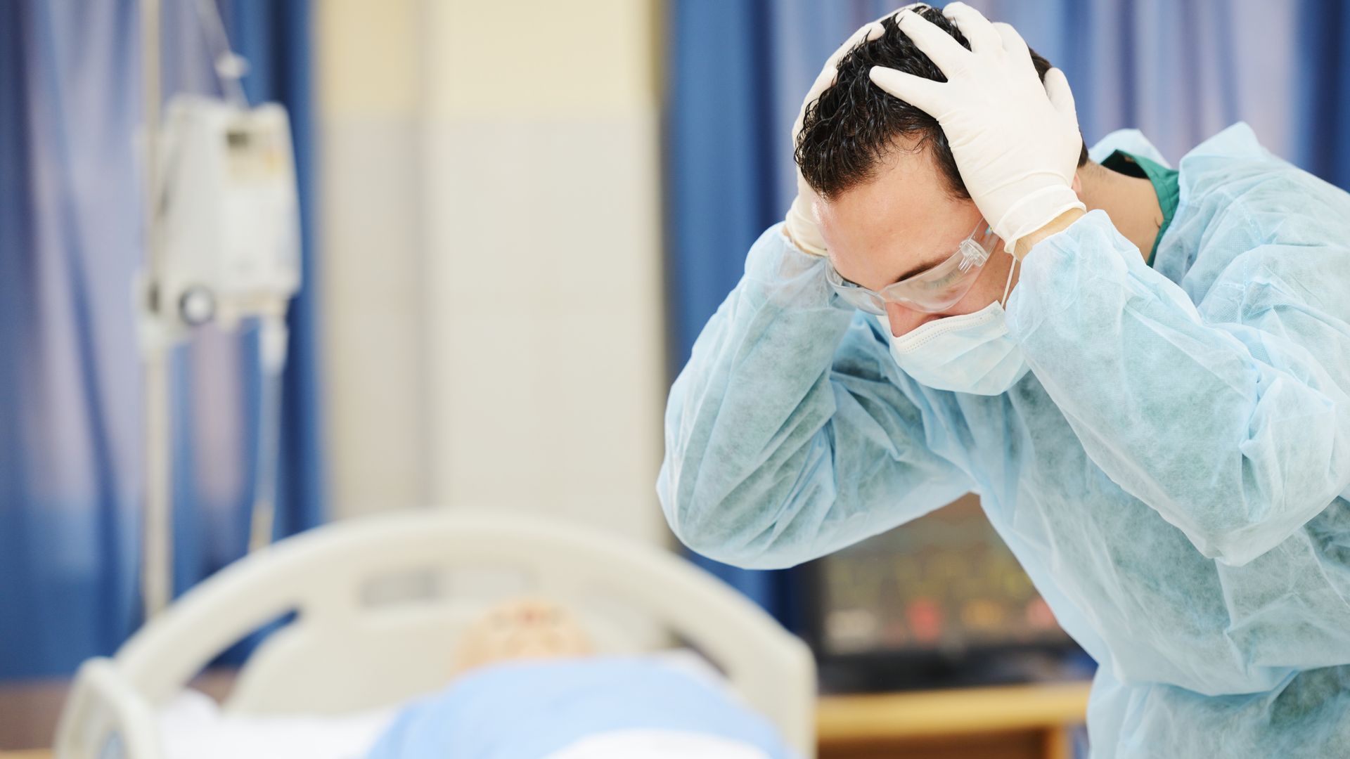 Doctor in scrubs holds head, stressed, near a patient's bed in a hospital room.