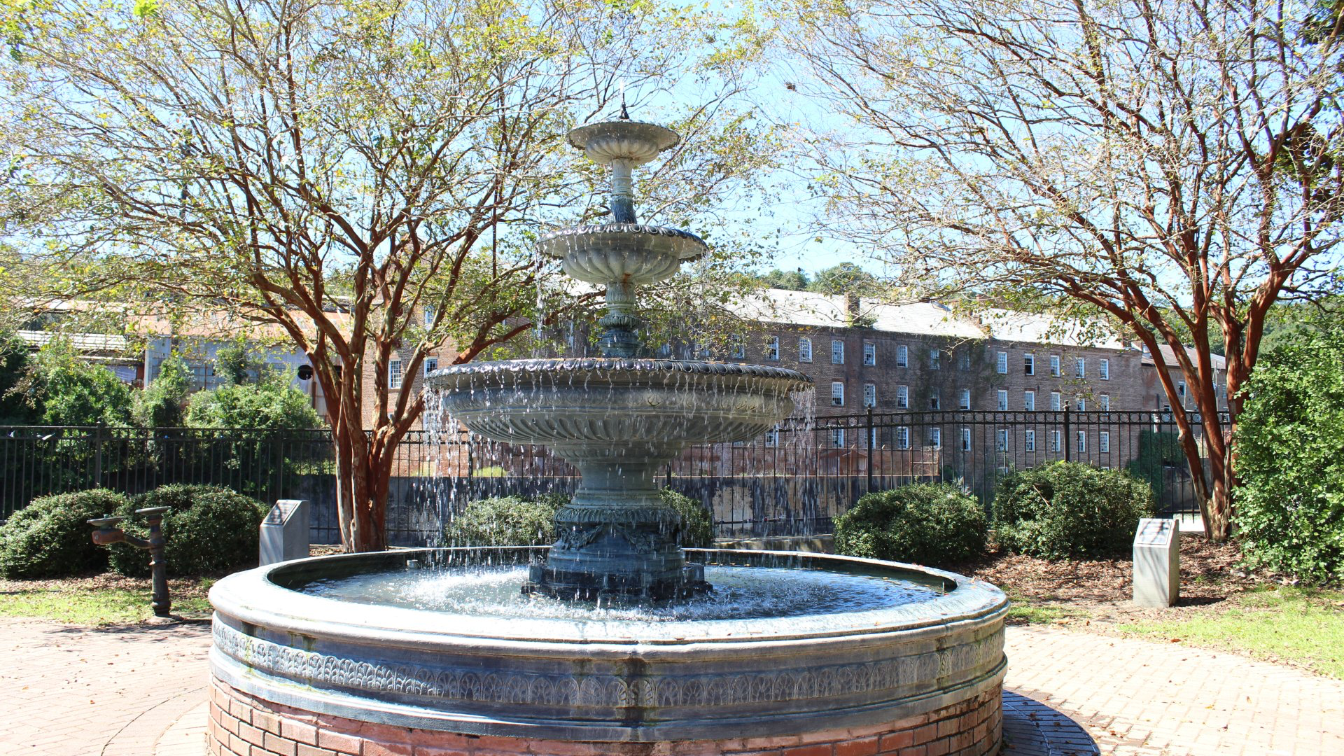 Three-tiered fountain in a brick and stone basin, surrounded by trees and shrubbery; buildings in the background.