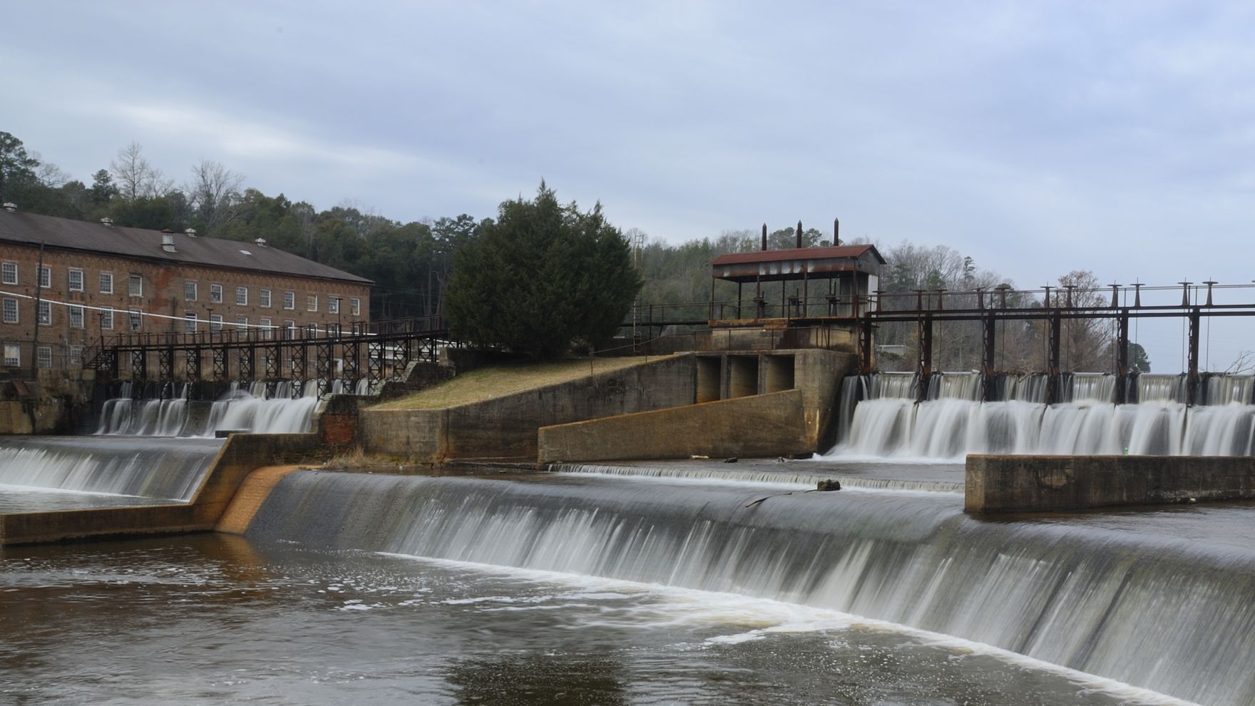 A low dam with water cascading over it; a brick building and a bridge are visible.