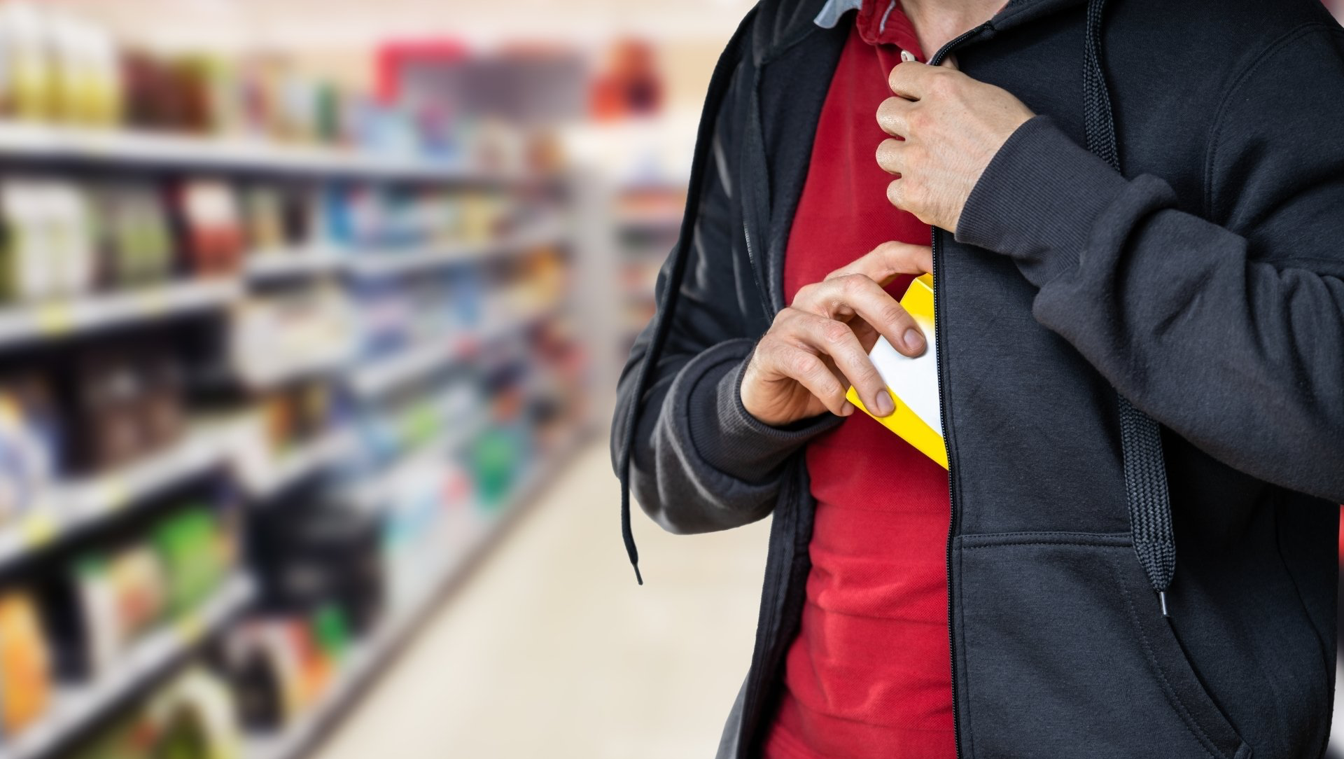 Person in a store shoplifting, putting item inside jacket. Blurred shelves in background.