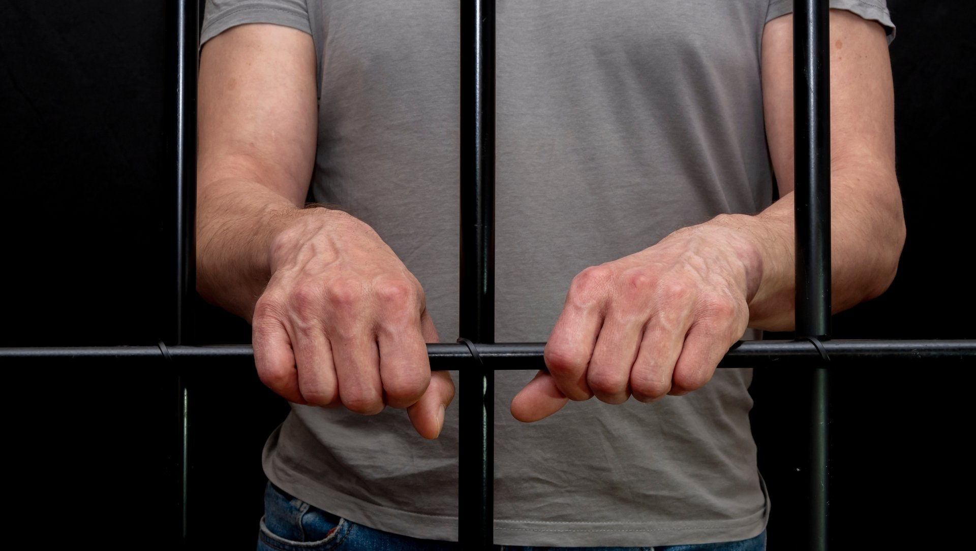 Person's hands gripping bars of a jail cell, wearing a gray shirt and jeans. Black background.