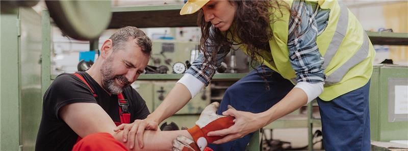 A person in a yellow vest examines another's injured leg in a factory setting. The injured person winces.