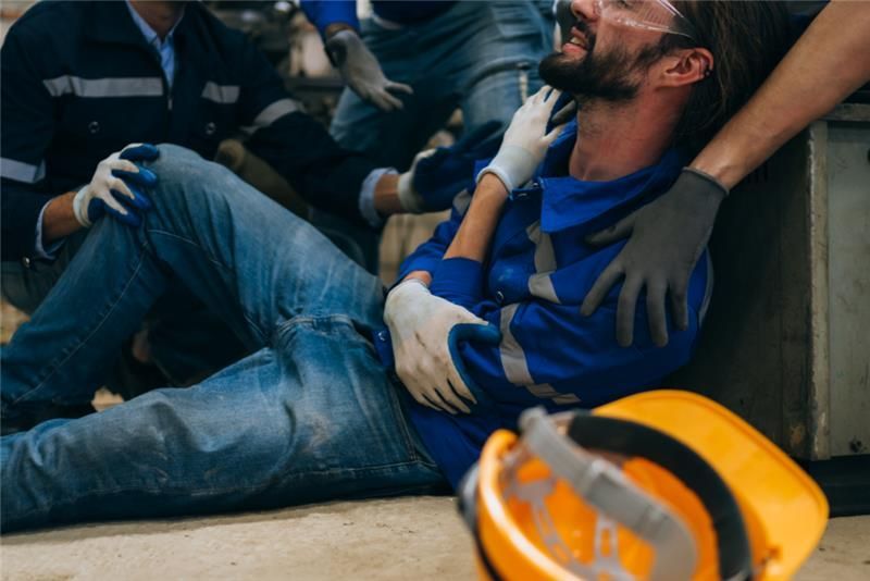 Man in blue jumpsuit injured, being helped by others in a work setting. Yellow hard hat on floor.