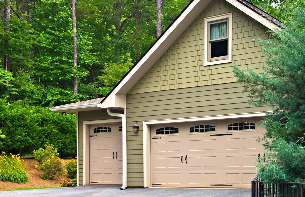 White house with green roof and attached garage beside a gravel driveway and grassy yard, with trees in back.