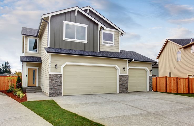 Modern two-story house with beige garage doors, gray siding, and a concrete driveway under a cloudy sky