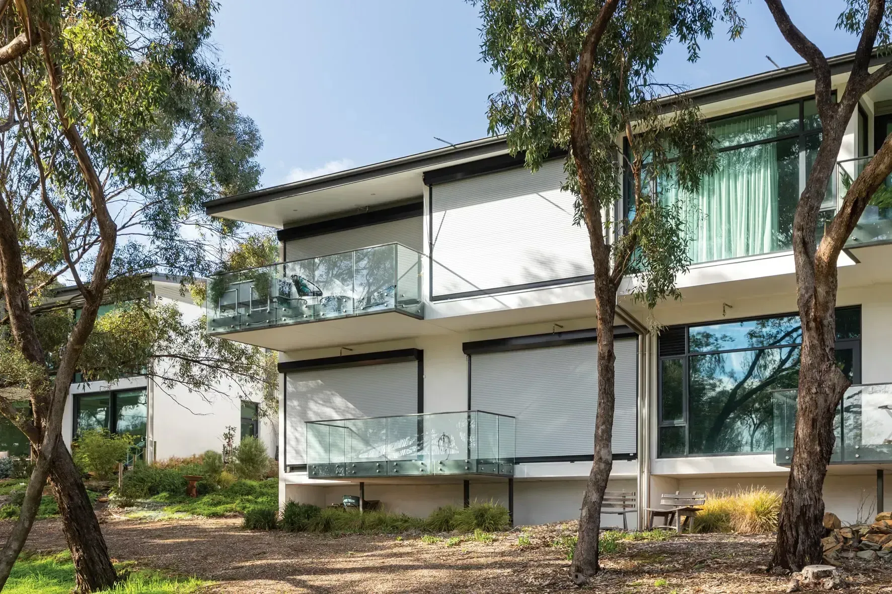 Modern two-story building with glass balconies and roller shutters. Trees frame the white exterior.