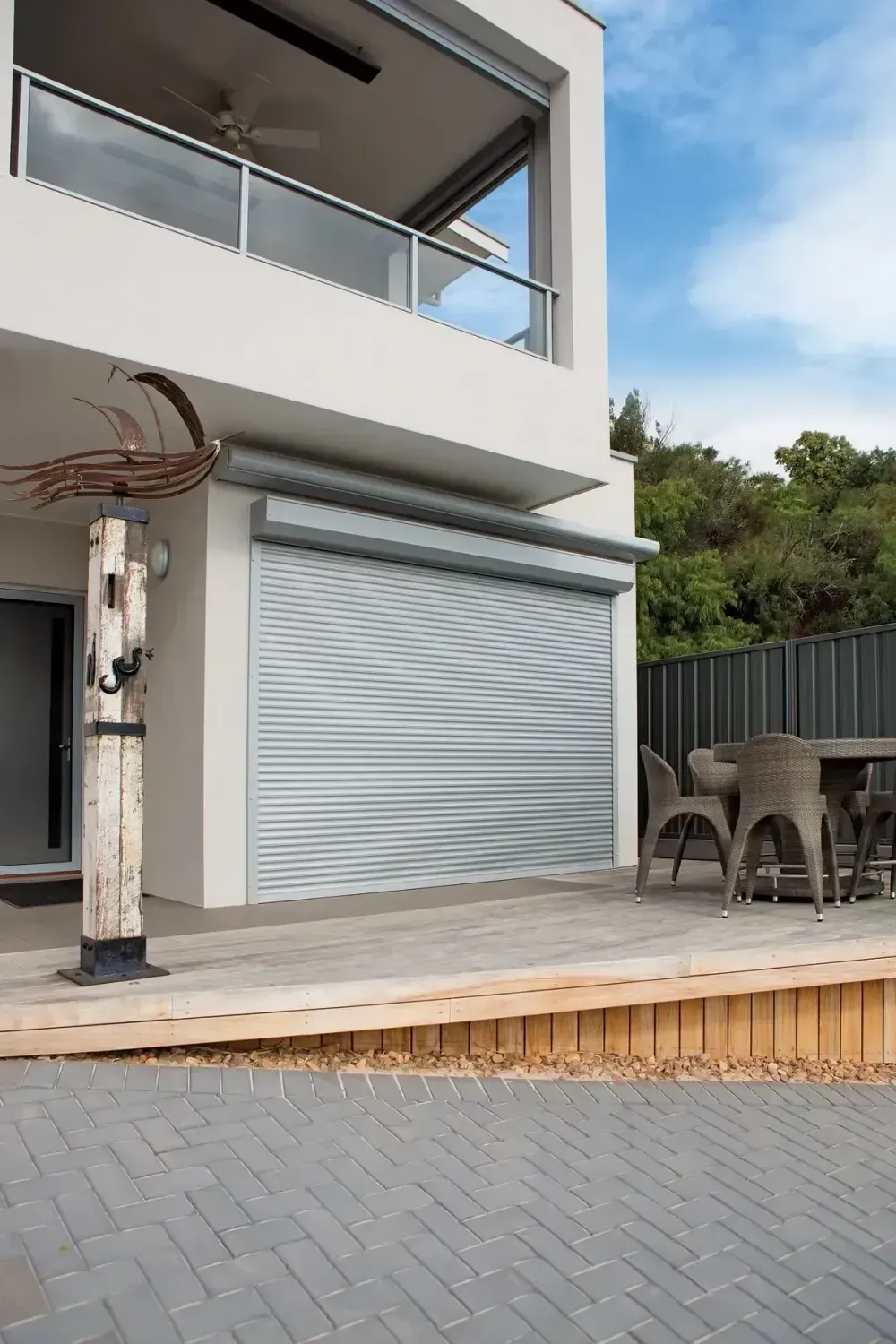 A modern white house with a gray roll-down shutter over a doorway, a wooden deck, and an outdoor table and chairs.