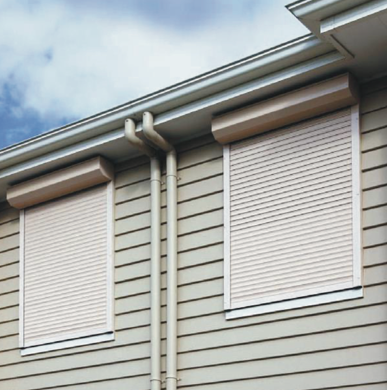 Two beige roll-down shutters on a building with horizontal siding, installed under a gutter.
