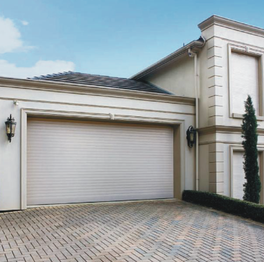 Beige roll-up garage door on a light-colored house with brick driveway and blue sky.