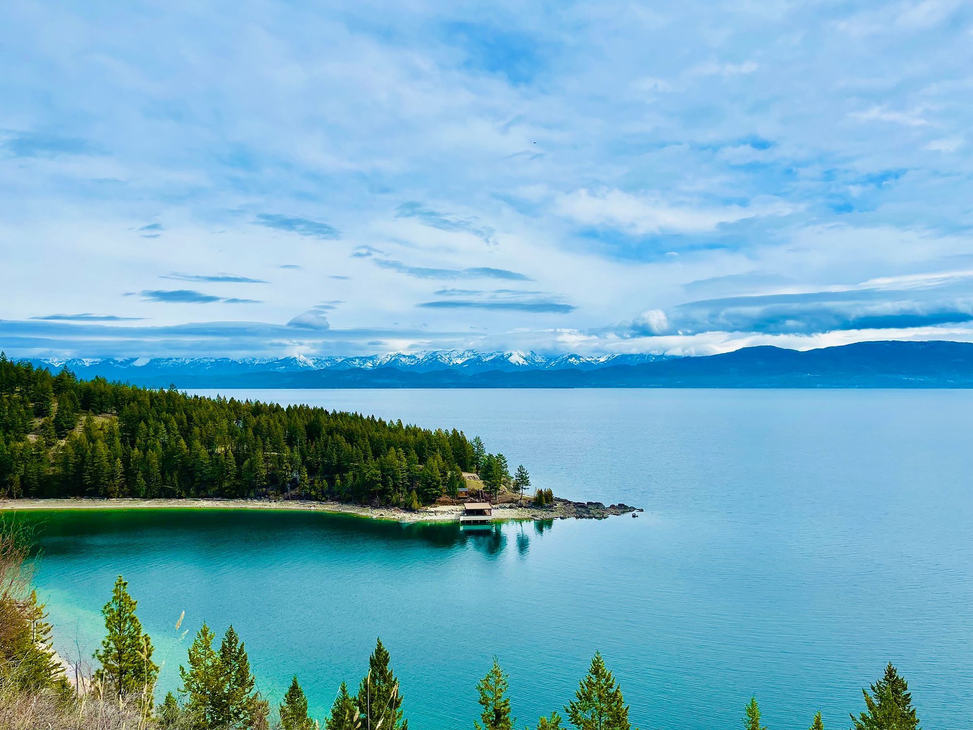 Blue lake with forested shoreline and distant mountains under a cloudy sky
