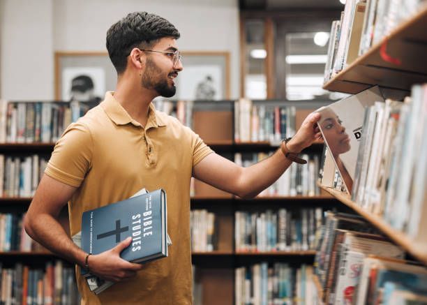 Man browsing books in a library, holding a notebook and reaching for a book on a shelf.