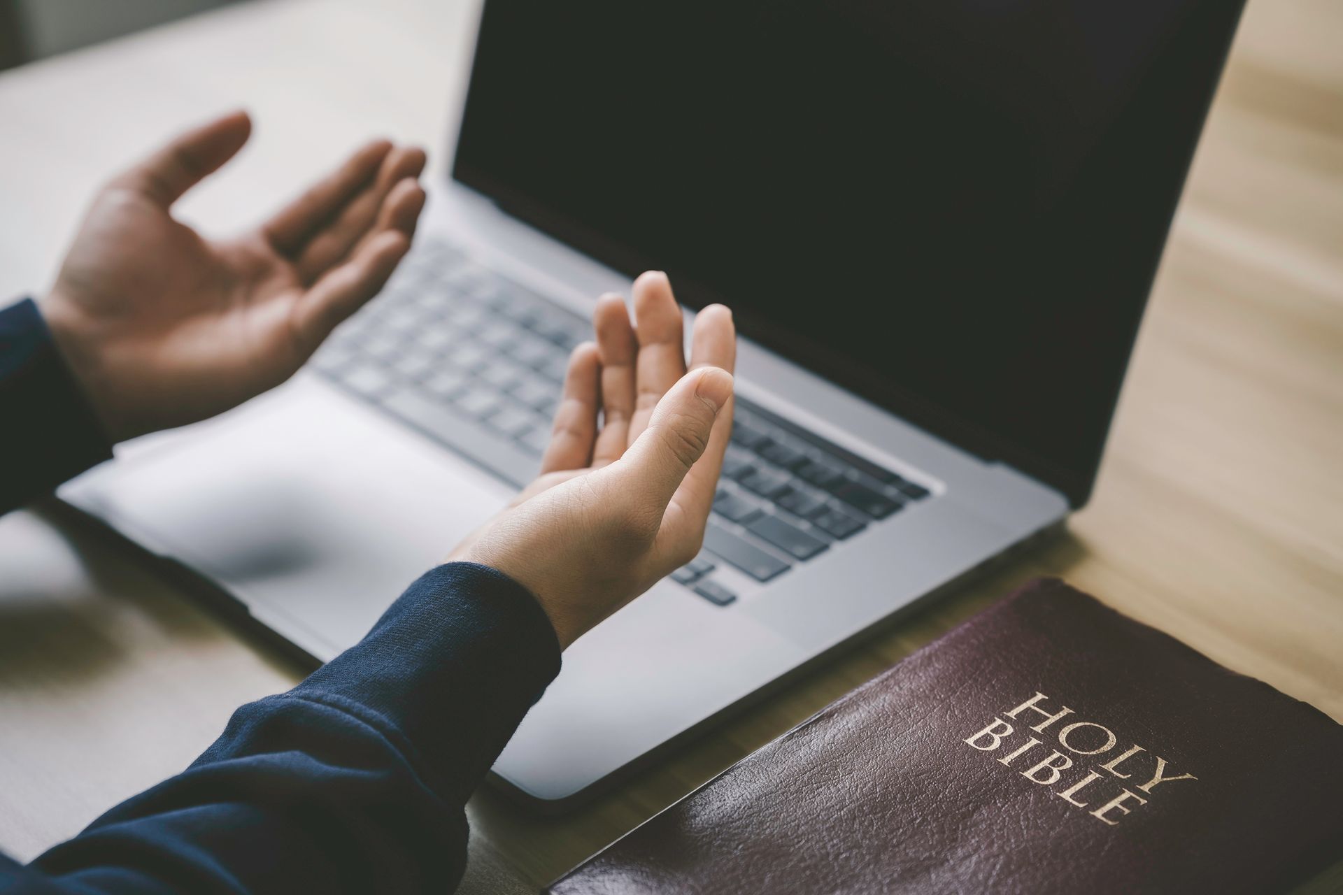 Hands raised in prayer beside an open laptop and a Bible on a table.