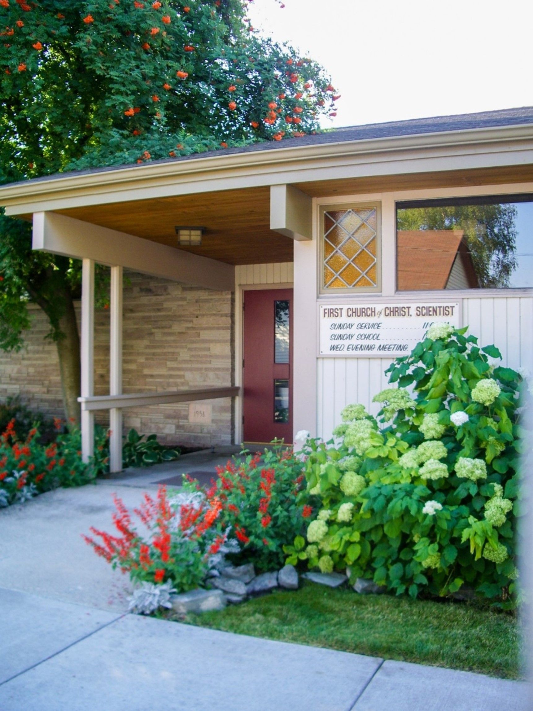 Modern house entrance with brick facade, covered porch, red door, and lush green landscaping
