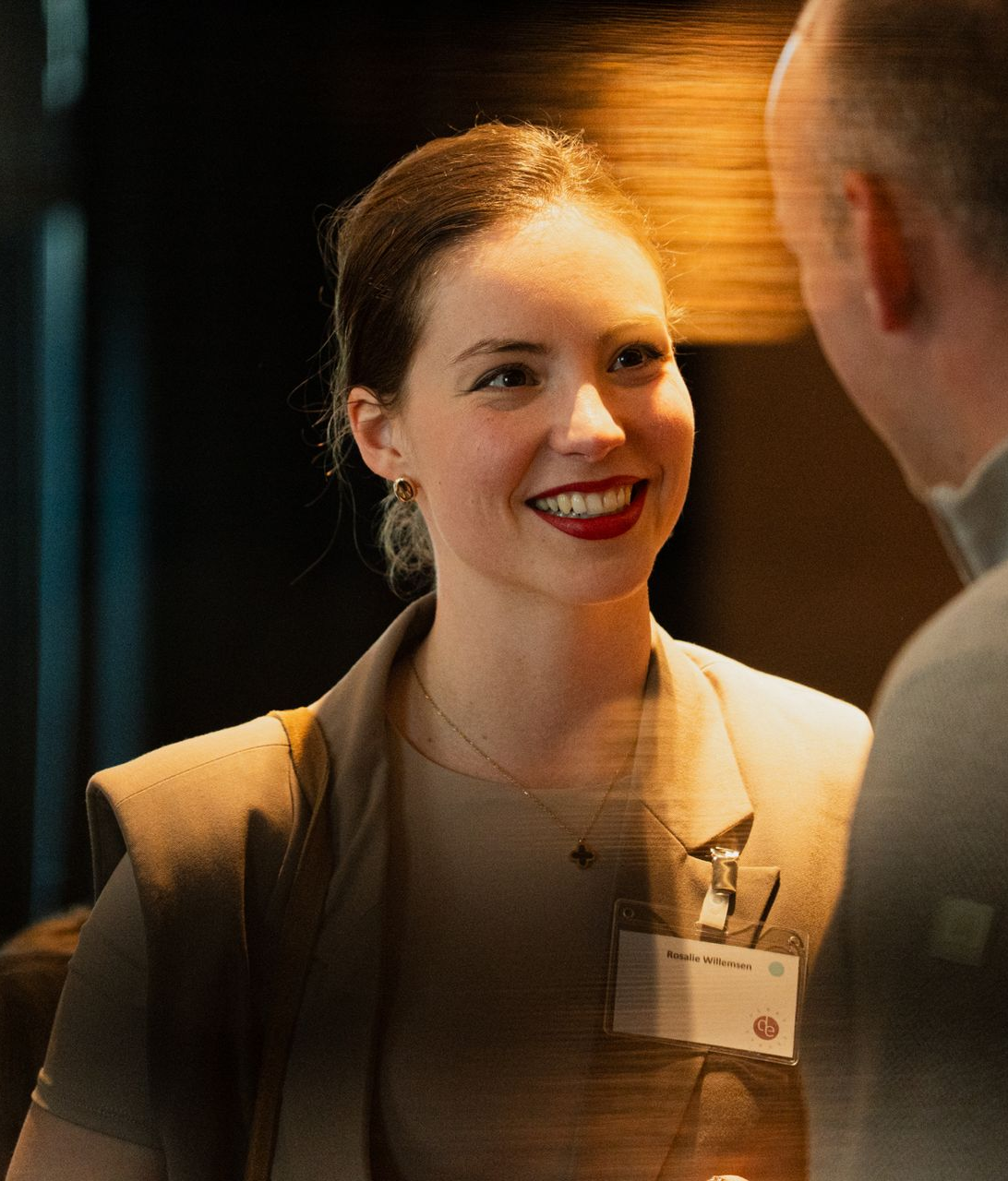 Vrouw in een beige blazer lacht en praat met iemand op een formeel evenement.