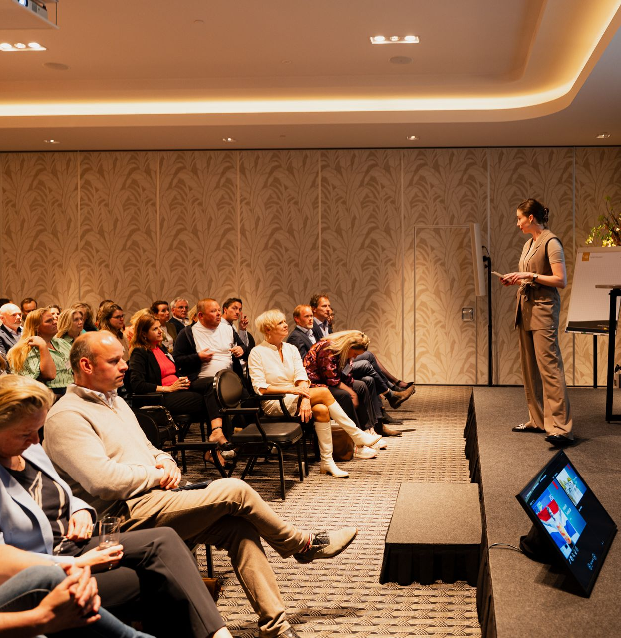 Het publiek zit in een conferentiezaal en luistert naar een spreker die op een podium staat.