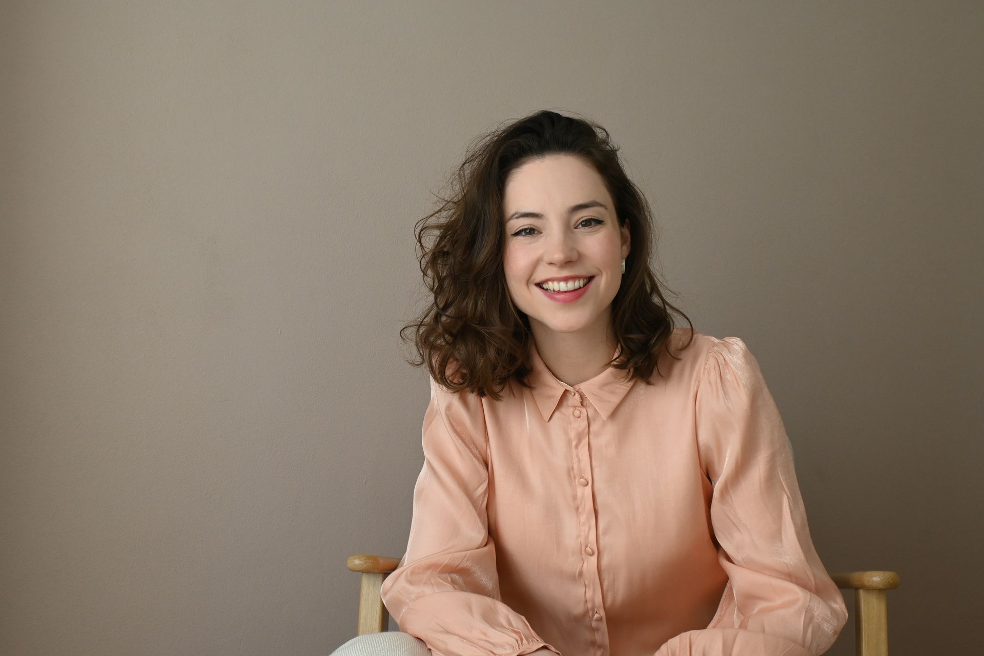 Lachende vrouw in een perzikkleurige blouse, zittend tegen een effen grijze achtergrond.