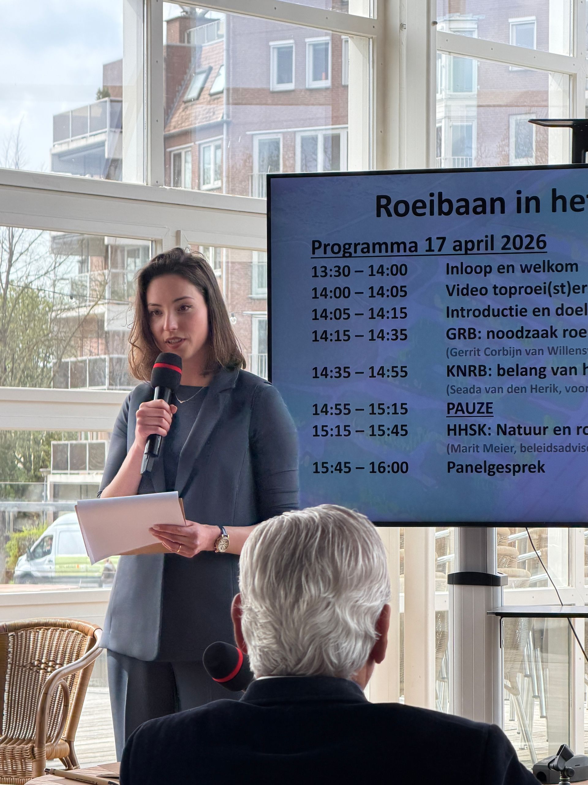 Een vrouw spreekt in een microfoon naast een scherm met het evenementenprogramma in een lichte, overdekte ruimte.