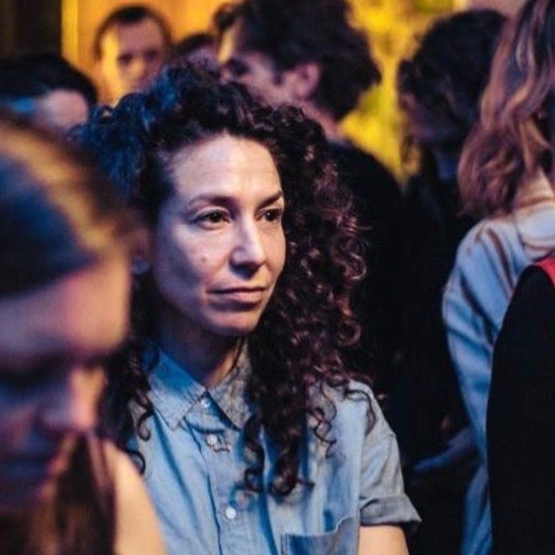 Een scène met een menigte, waarin een vrouw in een blauw shirt voor zich uit kijkt onder paarse en warme verlichting.