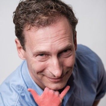 Een lachende man in een blauw shirt kijkt naar beneden, met een rode handpop bij zijn kin.