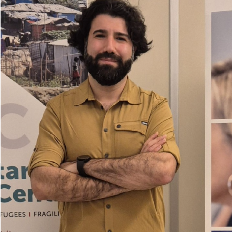 Een man staat met zijn armen over elkaar, gekleed in een mosterdkleurig shirt, voor een posterdisplay.