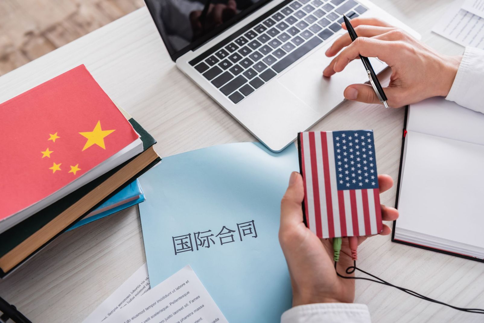 Hands holding US and China flag cards over a desk with laptop and business documents