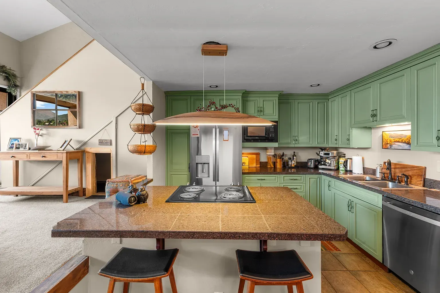 Green kitchen with island, pendant light, and stainless appliances in a bright open room