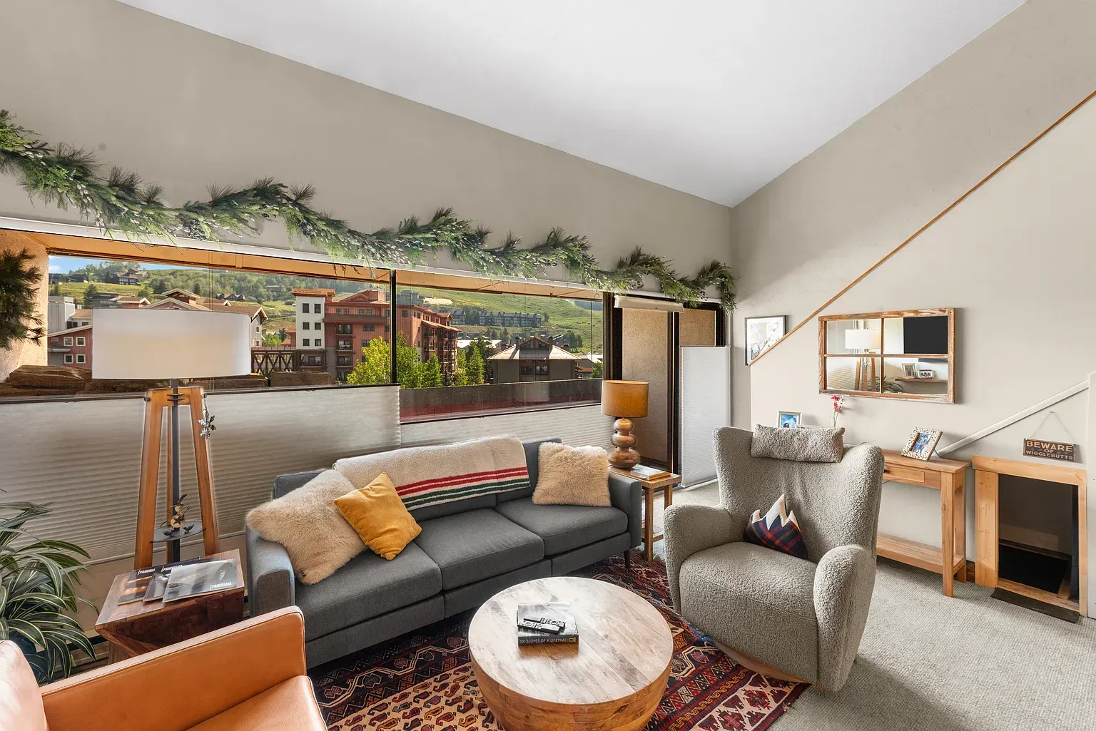 Cozy living room with gray sofa, patterned rug, armchairs, and a wooden desk beneath a garlanded window.