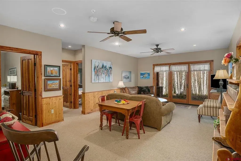Spacious beige living room with wood paneling, ceiling fans, dining table, fireplace, and large windows