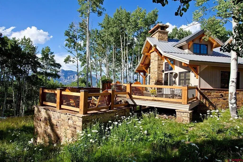 Wooden chalet with deck in a sunny forest clearing, surrounded by trees and wildflowers