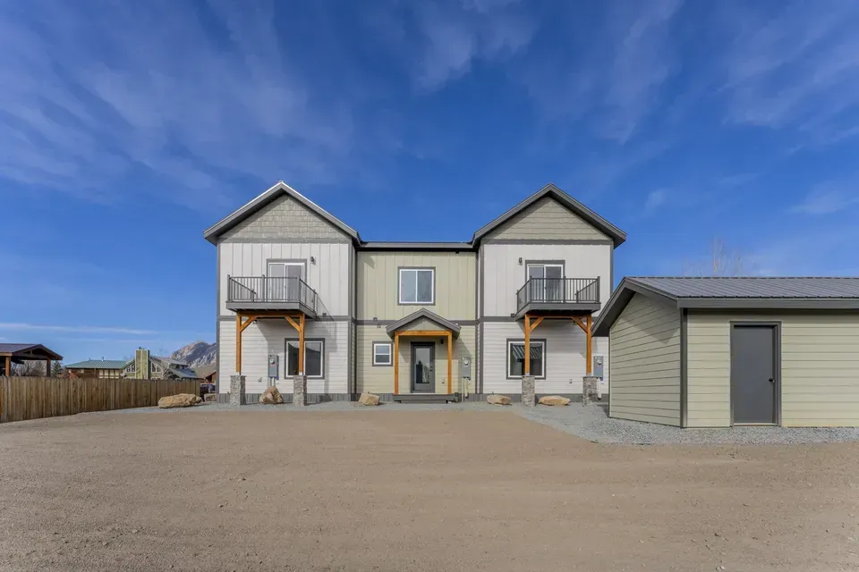 Two-story beige house with attached garage and gravel driveway under a blue sky