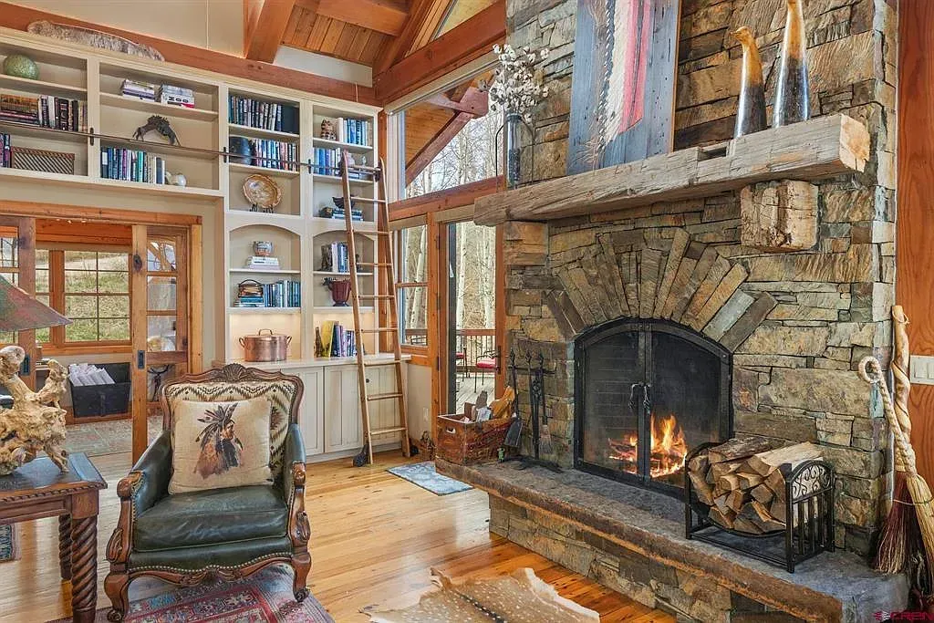 Rustic living room with stone fireplace, wood ceiling, and shelves of books and framed photos