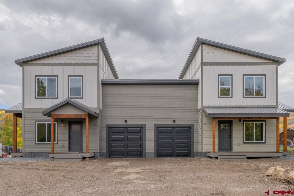 Modern gray duplex with two garage doors, front porches, and gabled roofs on a gravel lot