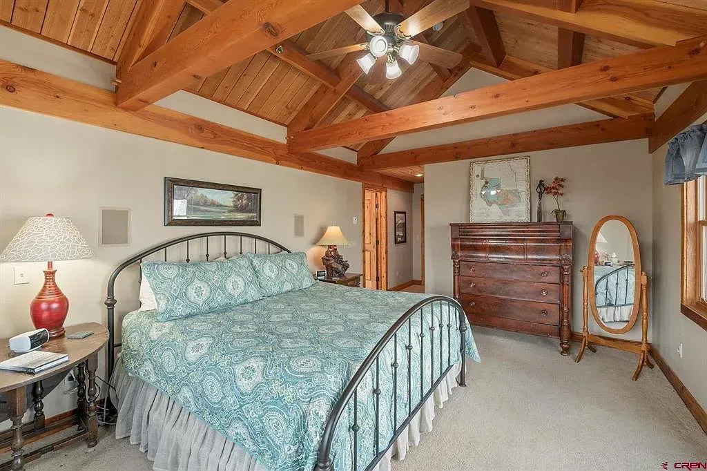 Rustic bedroom with wood-beamed ceiling, turquoise bedspread, dresser, and standing mirror