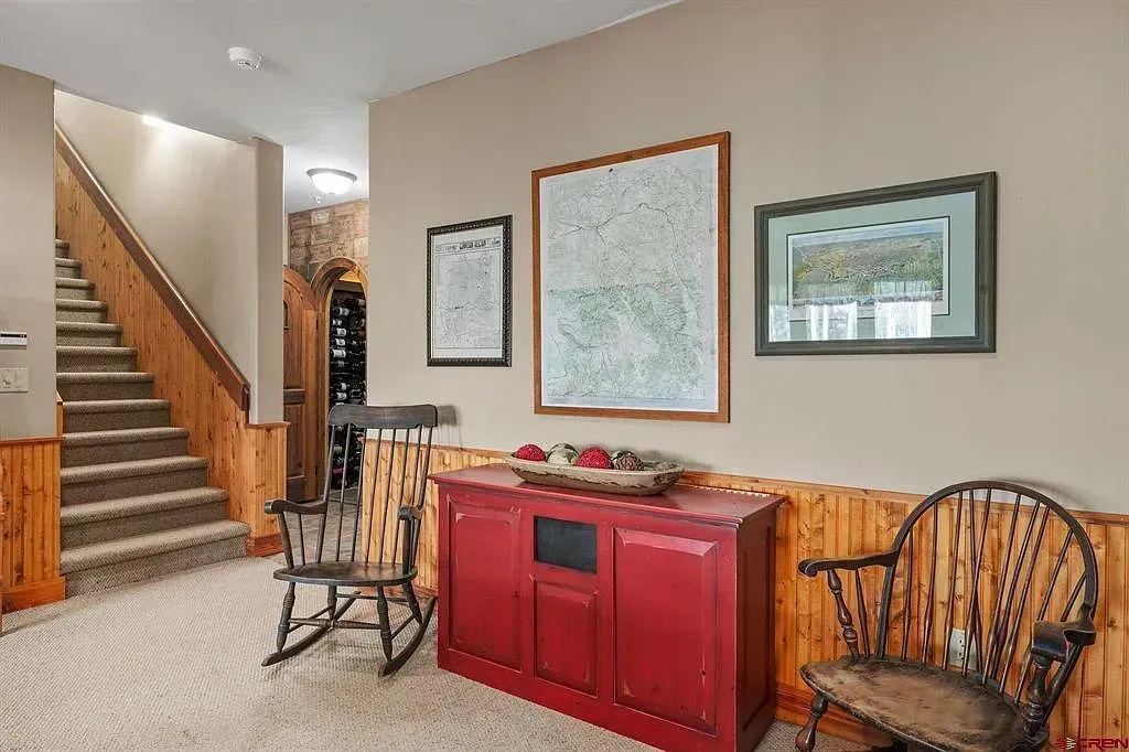 Living room with staircase, red cabinet, framed art, and two wooden chairs on carpet.