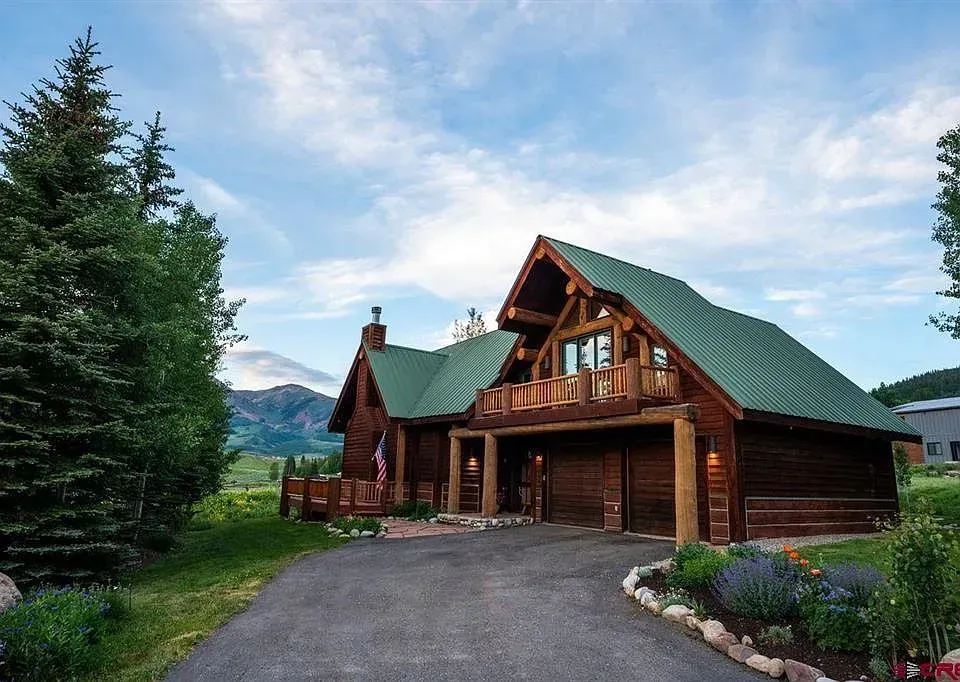 Rustic log cabin with green roof and garage, set beside tall pine trees under a blue sky.