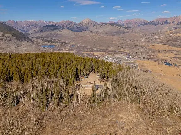 Mountain valley with autumn trees, a small cabin, and rugged peaks under a blue sky.