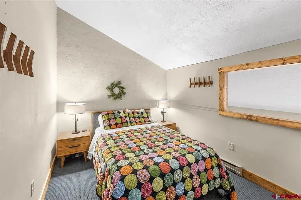 Small bedroom with colorful floral bedspread, white walls, wooden decor, and a wall mirror.