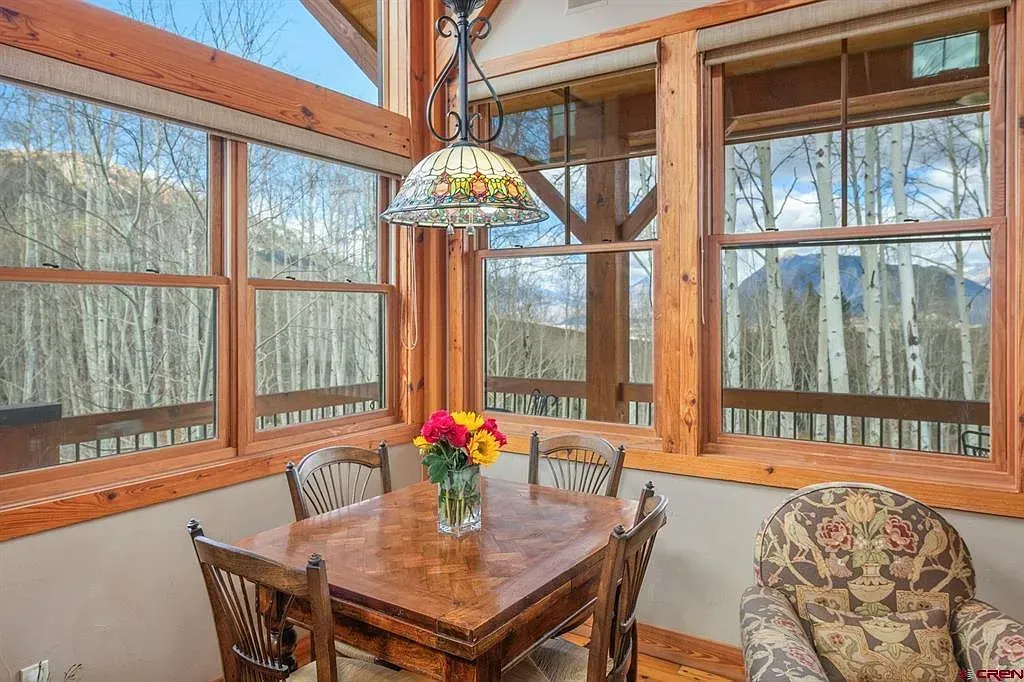 Sunroom dining area with wood table, floral lamp, and large windows overlooking trees