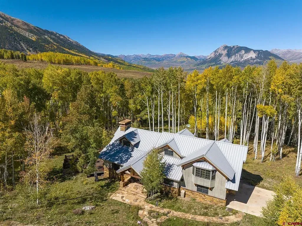 A rustic cabin with a metal roof sits in a forest of yellow aspens beneath mountains.
