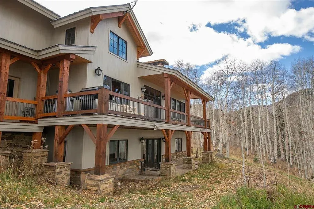 Two-story house with wooden balconies beside a leafless forest under a cloudy sky