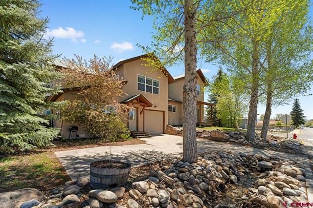 Two-story beige house with stone landscaping and tall trees on a sunny day