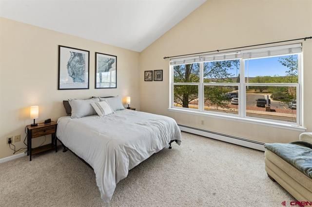Bright bedroom with a large window, white bed, and beige carpet, overlooking a sunny landscape.