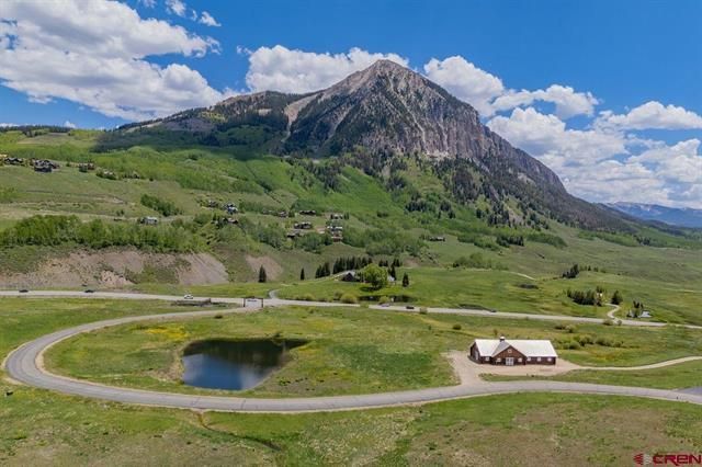 Mountain landscape with green meadow, winding road, small pond, and cabin beneath a rocky peak under cloudy blue sky